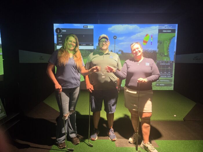 Two women and a man holding golf clubs and posing in front of a golfing screen.