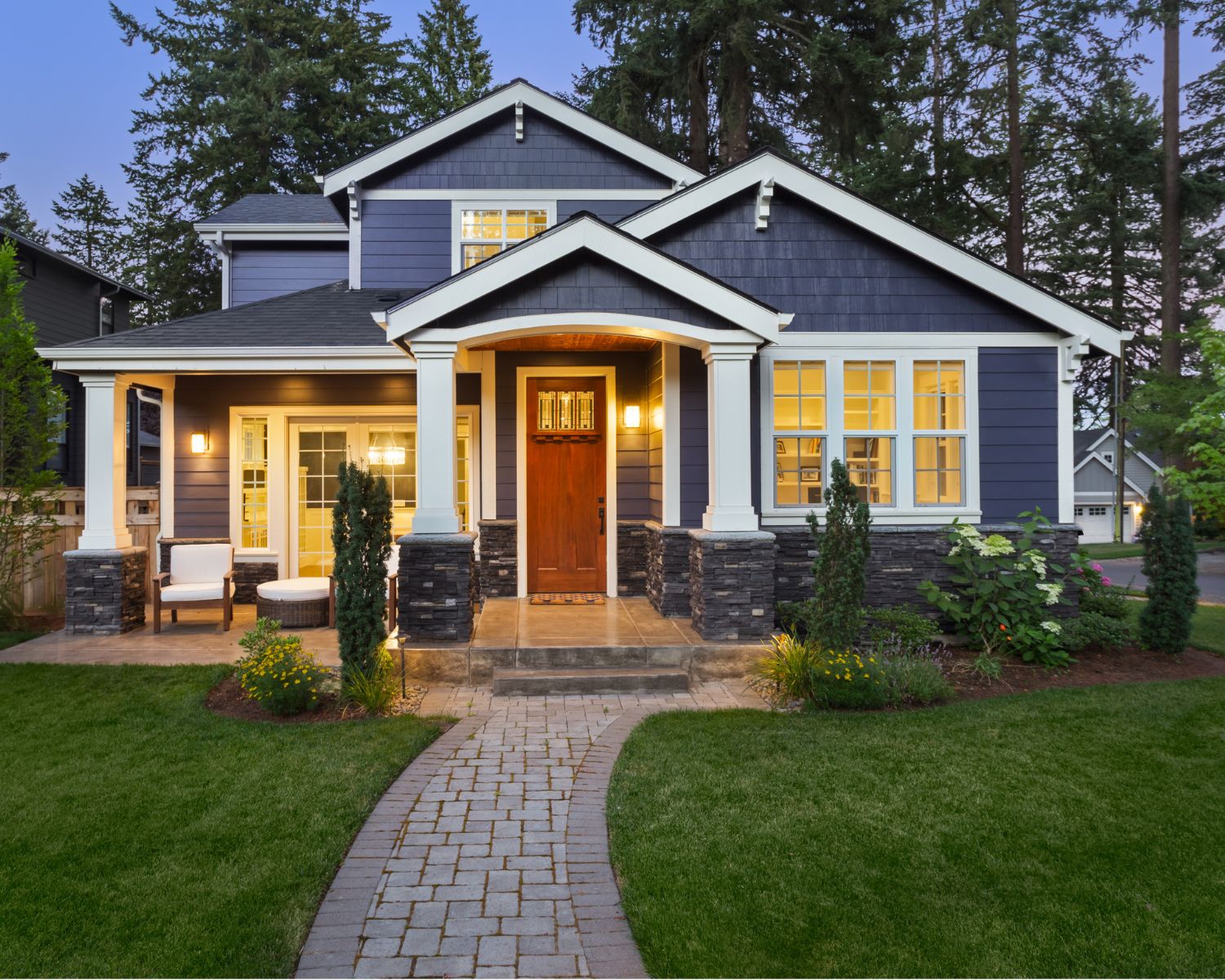 Medium‑to‑large blue house at sunset with warm interior lights glowing through the windows; sky shows orange and pink tones, creating a cozy, welcoming atmosphere.