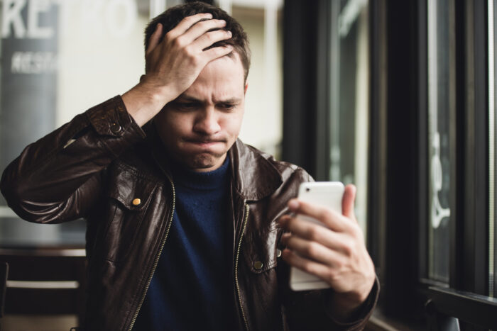 Closeup portrait, stressed young man in purple sweater, shocked surprised, horrified disturbed, by what he sees on his cell phone, isolated indoors background