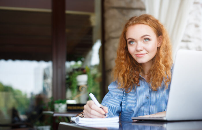 Young red-headed lady sitting outside at a table writing in a spiral journal with laptop opened next to her.